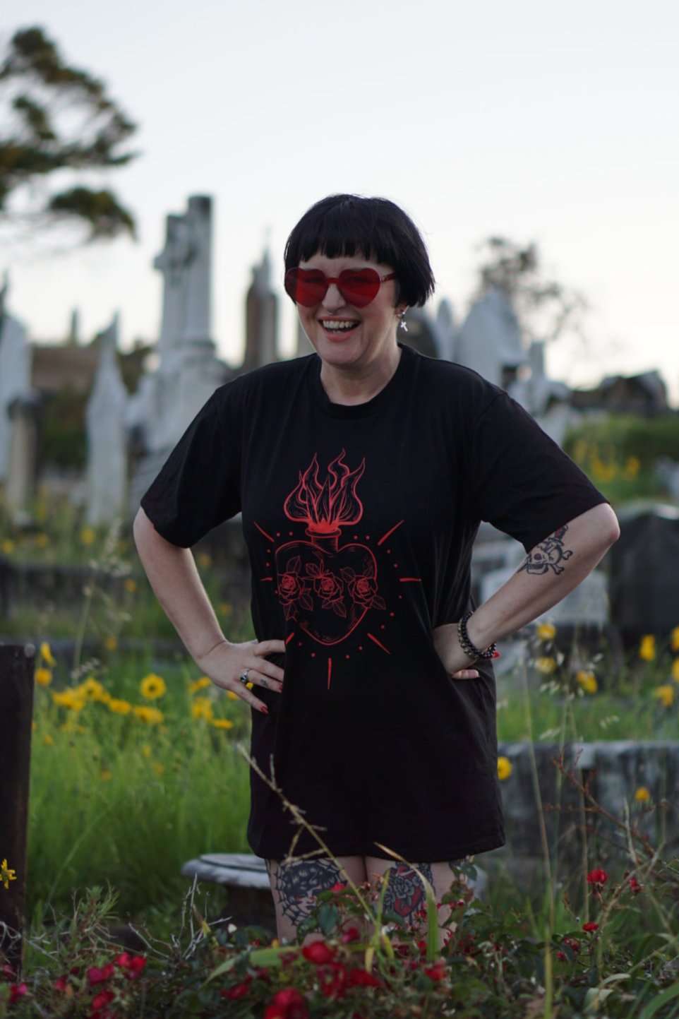 Smiling model in Queen of Hearts Xtra Long Tee/Dress with red sacred heart design, styled as oversized tee in a floral-filled cemetery scene.