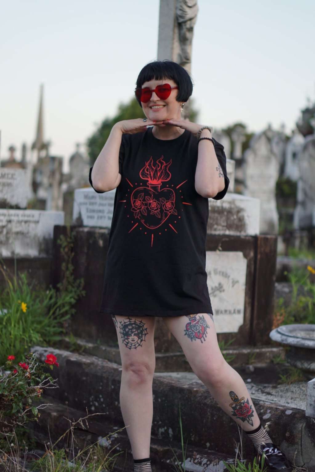 Model poses playfully in Queen of Hearts Xtra Long Tee/Dress, featuring bold red sacred heart print, styled as a t-shirt dress among tombstones.
