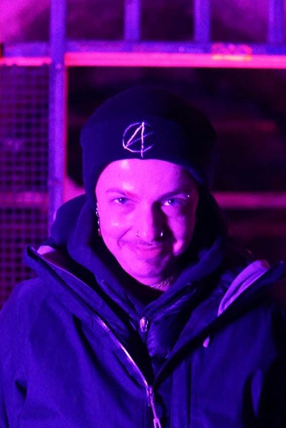 Non binary person wearing black beanie with white embroided A symbol on the front (anarchy)
