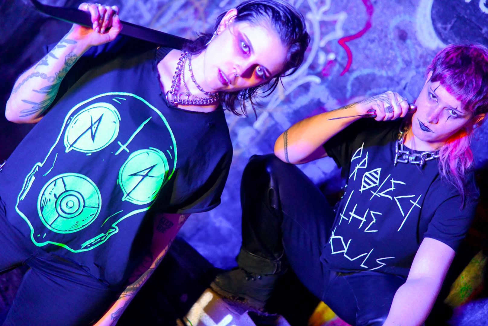 Post apocalyptic science - Non binary person wearing a black tshirt with a large toxic green gas mask & trans person wearing a black tshirt with protect the ghouls on the front in gothic font. Both people are holding weapons and wearing gothic makeup