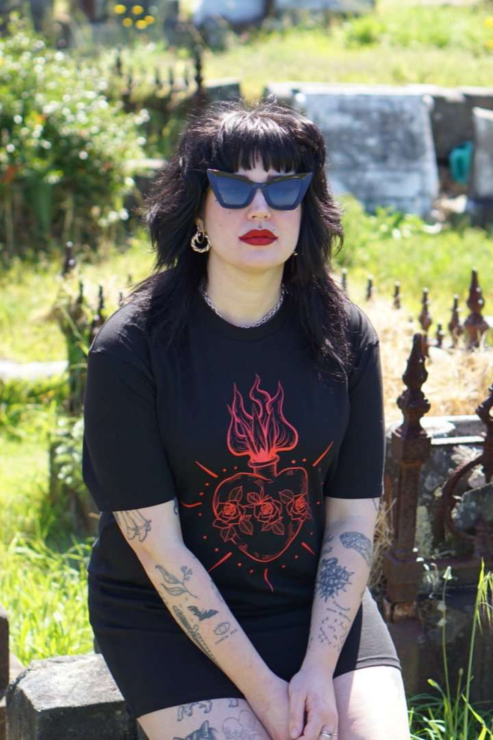 Model wearing Queen of Hearts Xtra Long Tee/Dress in black with red sacred heart graphic, styled as a dress in a gothic cemetery setting.