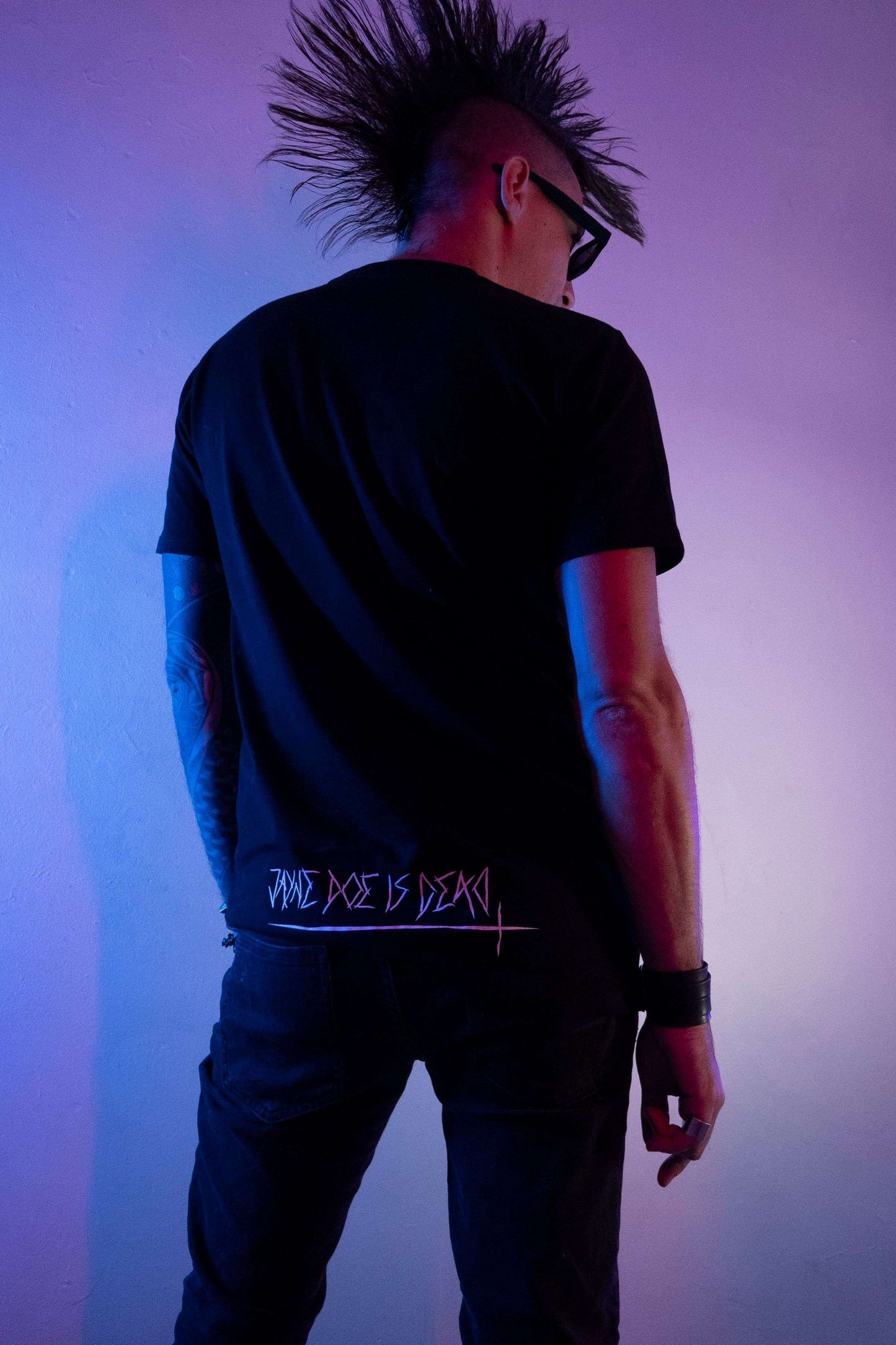 Tall guy with huge black mohawk wearing a black long tshirt with jayne doe is dead print along the bottom hem