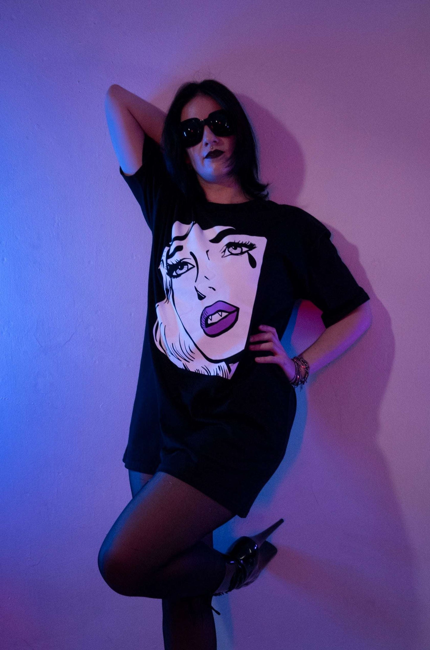 Tall curvy gothic girl with black stockins & patent leather pleasers with a black long tshirt with an extra large white pop art woman's face crying one tear with fangs & purple lips.