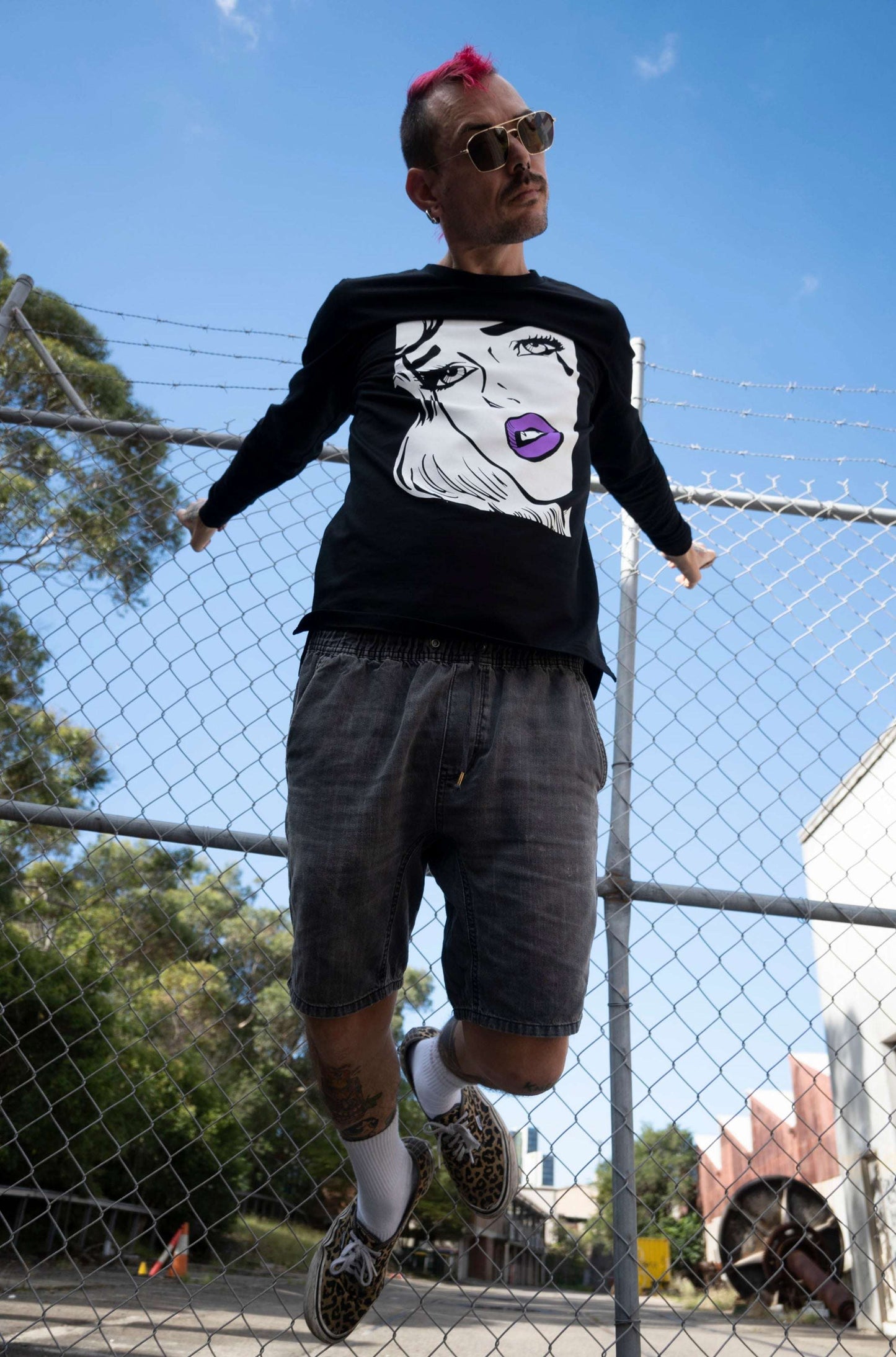 Guy hanging from wire fence like a vampire wearing a long sleeve pop art vampire tshirt in black with a white face and purple lips & fangs, sleeve print is jayne doe is dead logo