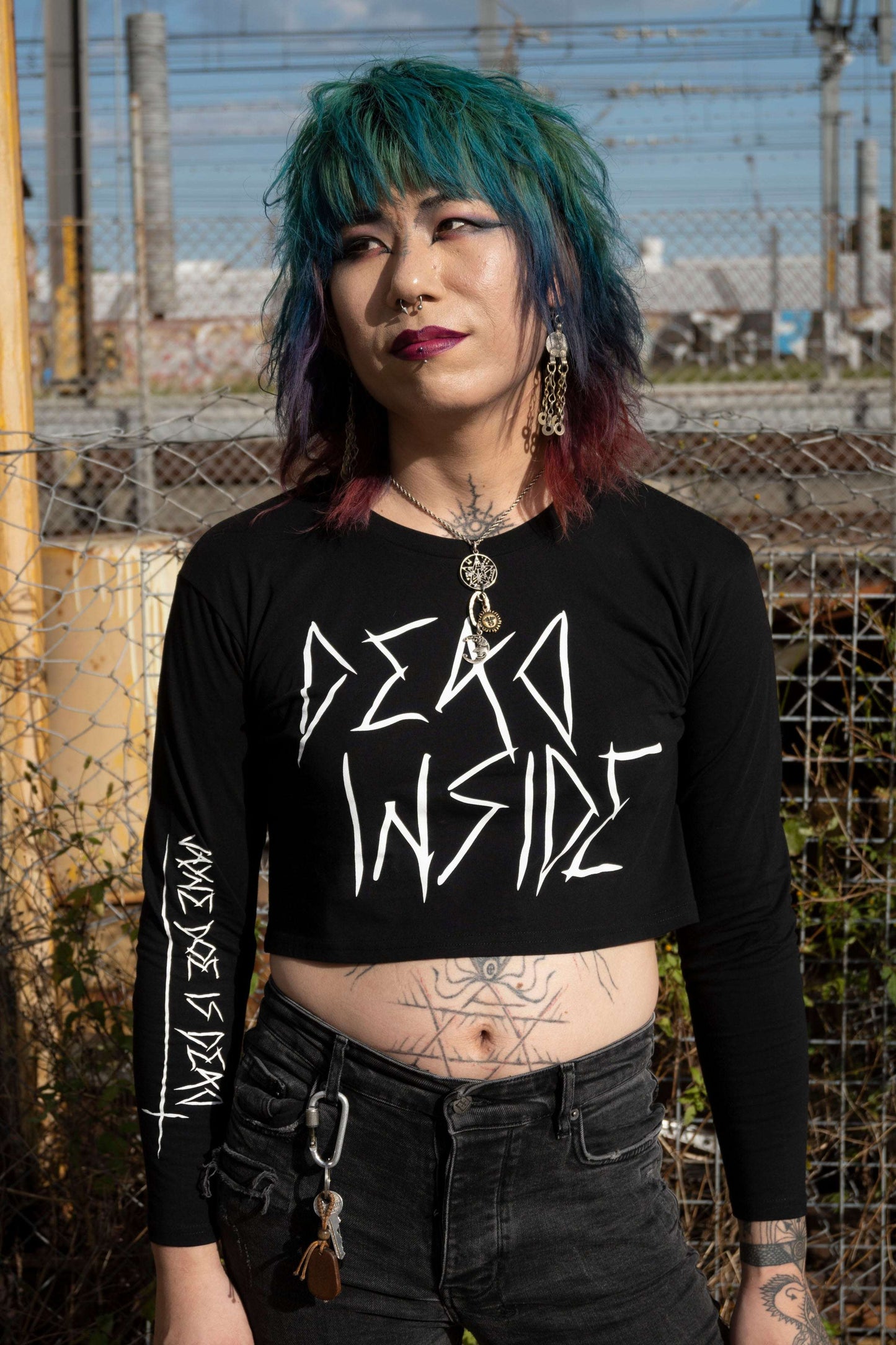 Goth trans girl wearing a long sleeve black cropped tshirt with the words dead inside printed on the front in white gothic font. The arm has a jayne doe is dead