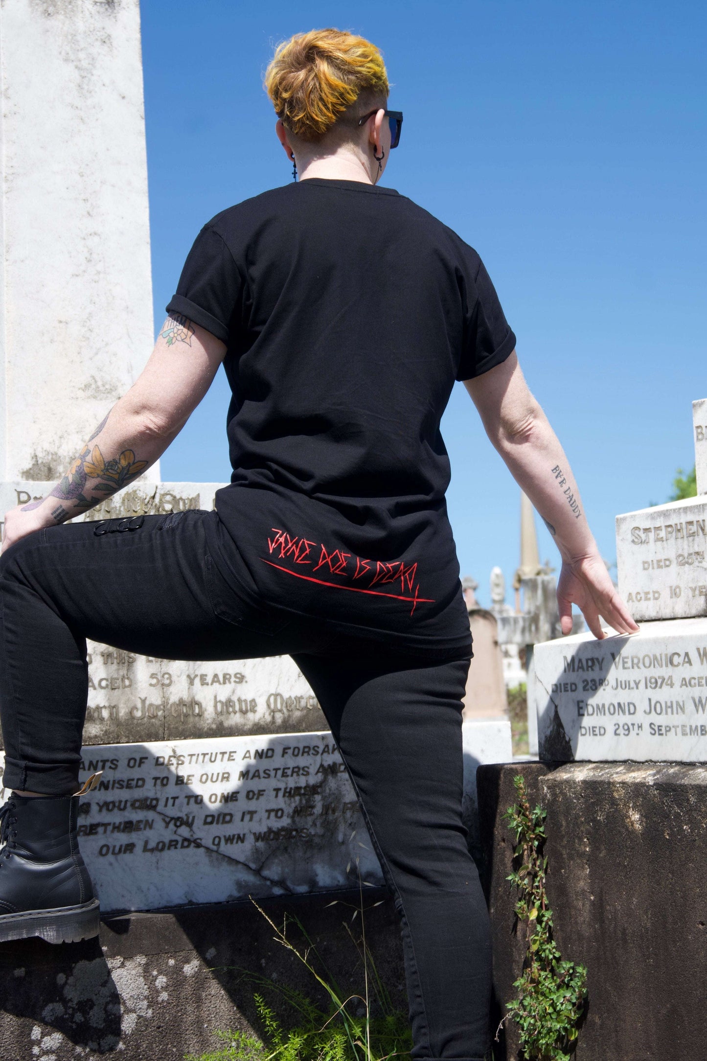 Queen of Hearts Tee in black with red 'Jayne Doe is Dead' text on lower back, styled with black jeans and boots in a gothic cemetery setting.
