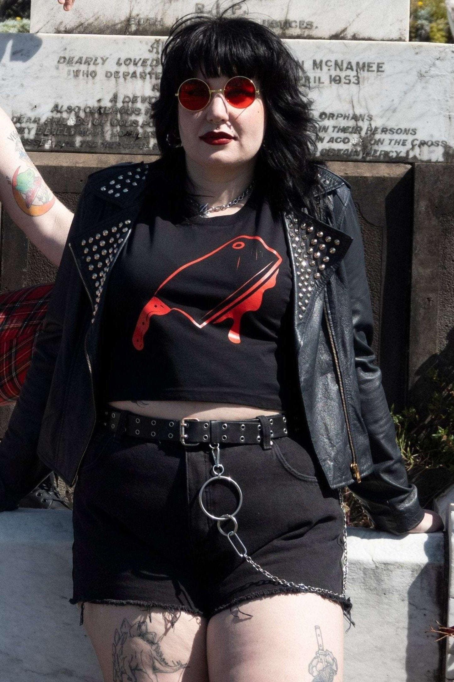Off with their Heads Cropped Tank featuring a red cleaver graphic, styled with black shorts and studded leather jacket in a gothic cemetery setting.