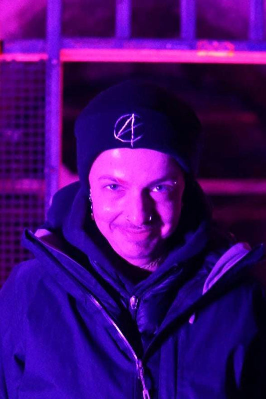 Non binary person wearing black beanie with white embroided A symbol on the front (anarchy)