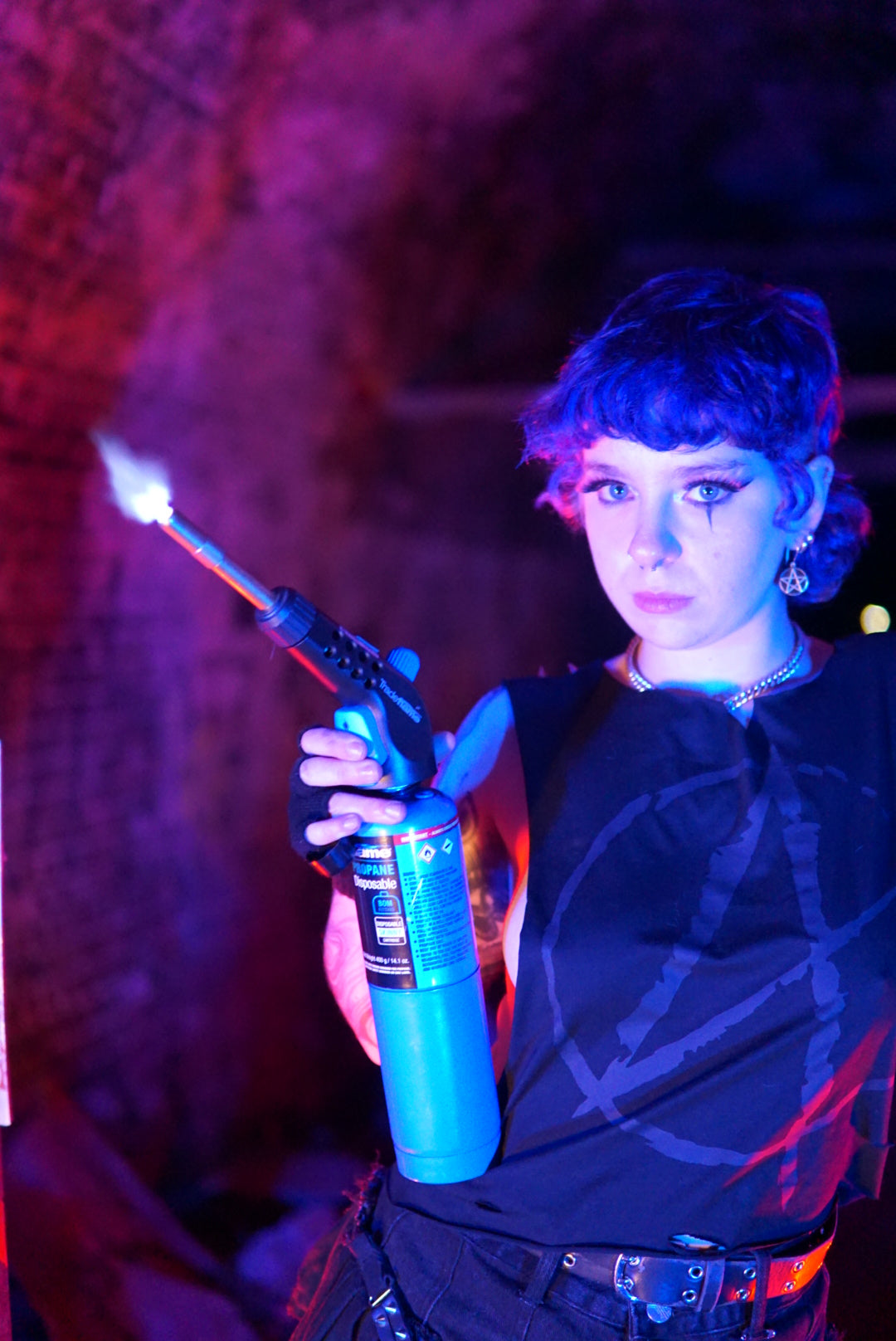 Non binary person wearing a size small Anarchy Cropped Tank with a goth black A print on the front. Holding a blow torch in a post apocalyptic scene