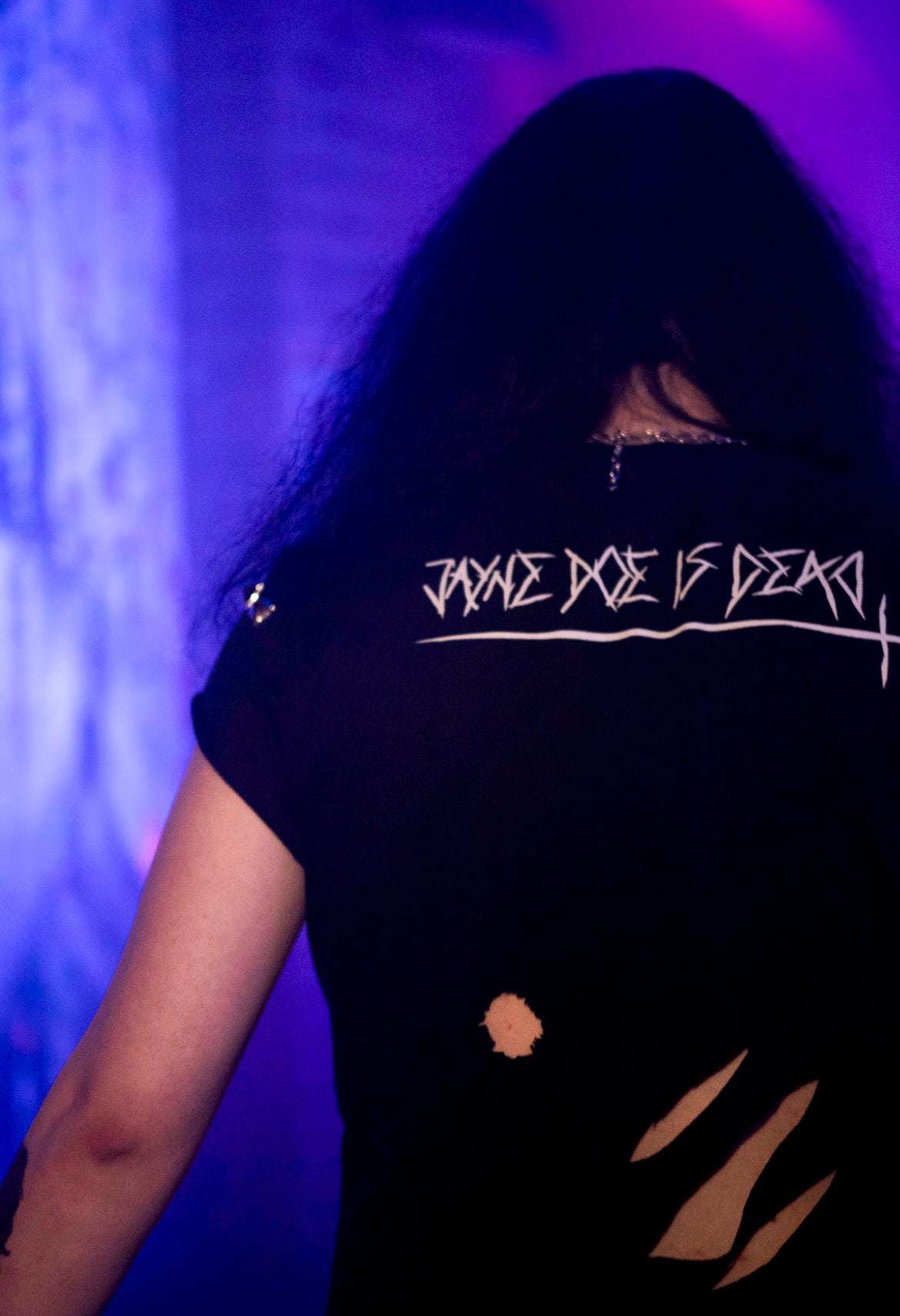Goth guy with long hair wearing a black cropped tshirt with a white Jayne doe is dead logo in goth font on the back