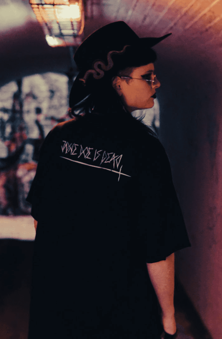 Post apocalyptic style black tshirt with white jayne doe is dead - Desolate in black, featuring bold graphic print against graffiti wall